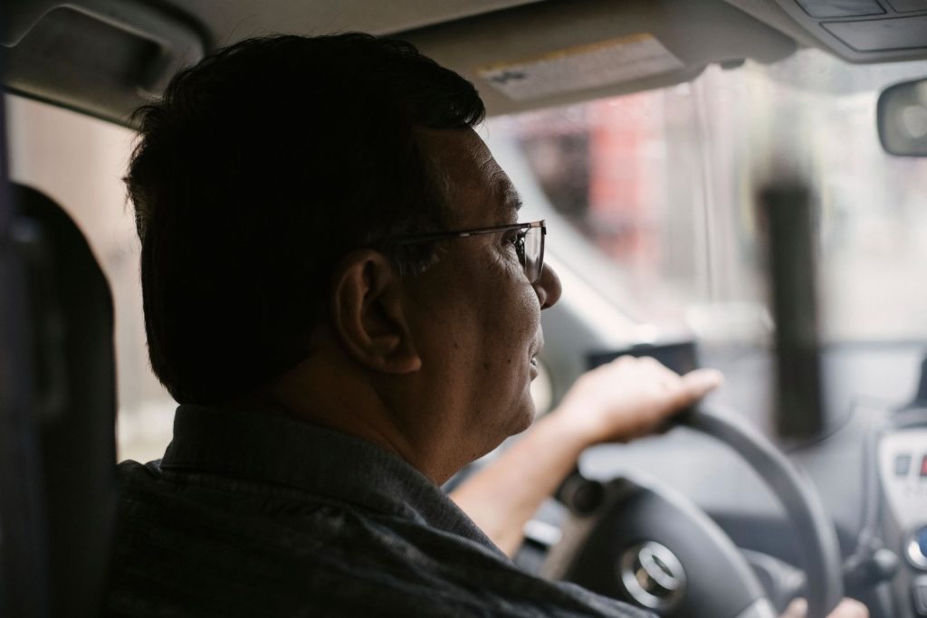眼鏡をかけたシニアの男性が、車のハンドルを握って前方を見ながら運転している様子